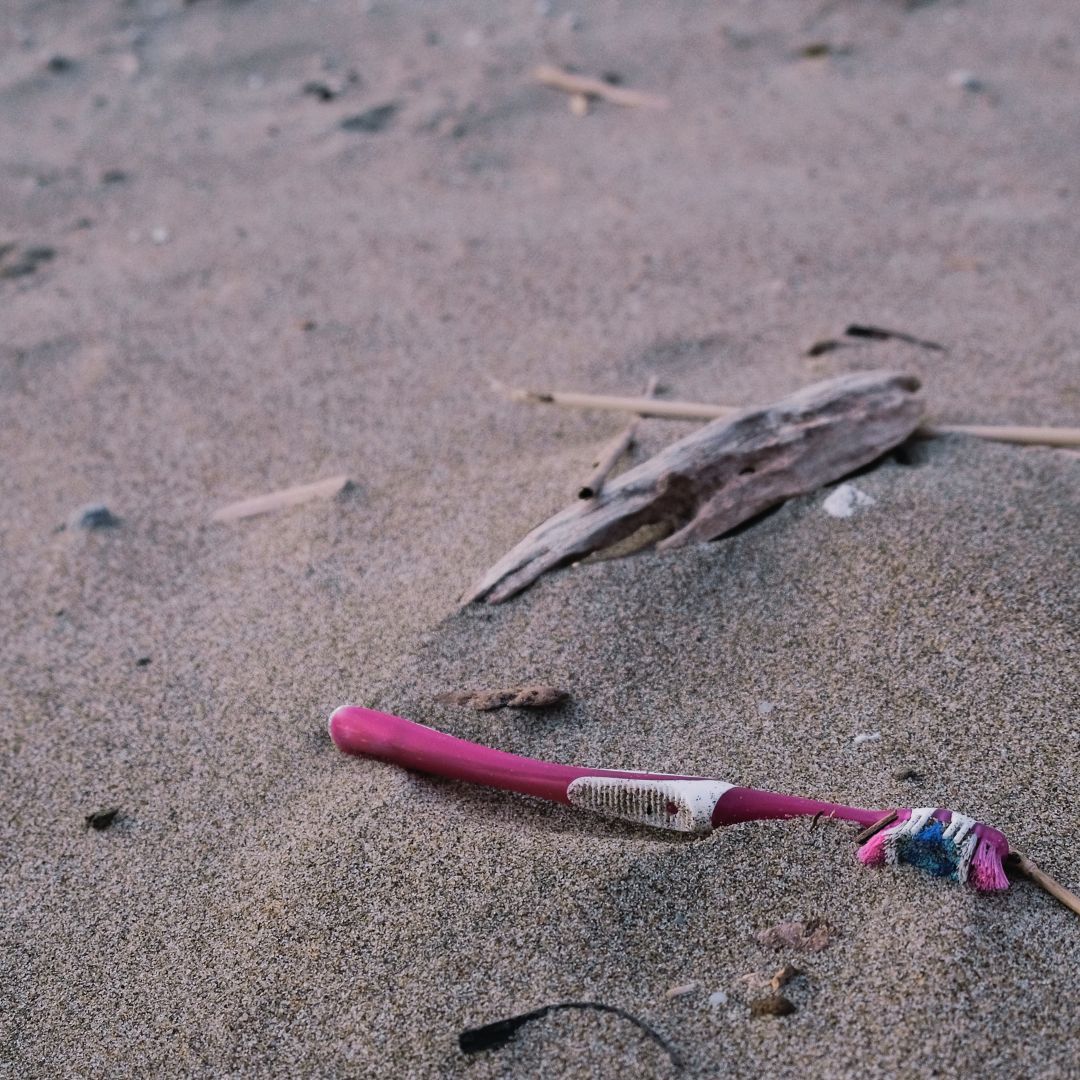 A broken pink plastic toothbrush lying on sand beside a piece of driftwood.